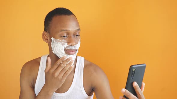 Confident African-American Man Applies Shaving Foam Using His Smartphone Camera As a Mirror