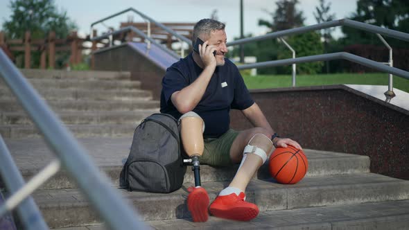 Wide Shot Motivated Positive Male Amputee Talking on Phone Sitting on Park Stairs with Basketball