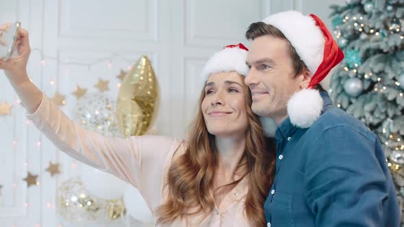 Smiling Couple Making Selfie in Santa Hats Near Christmas Tree.