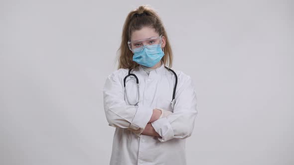 Little Nurse Doctor in Coronavirus Face Mask Medical Gown and Eyeglasses Crossing Hands Posing at