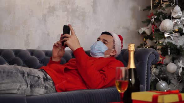 Man in Medical Mask and Santa Hat Browses Smartphone, Lying on Couch in Room. Young Guy in