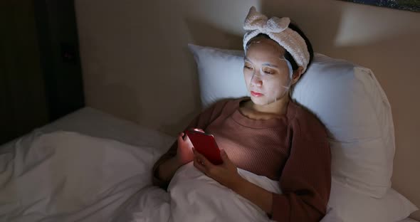 Woman use of mobile phone and apply the face mask on bed at night