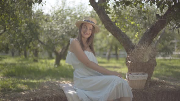 Cute Young Woman in Straw Hat and Long White Dress Looking at the Camera Smiling Sitting