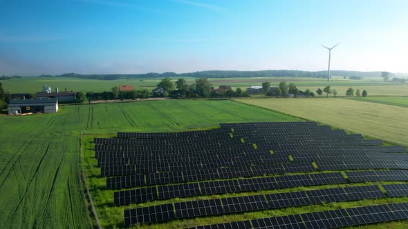 Scenic View Of Solar Panels On Green Field With Wind Turbine In Background - aerial drone shot