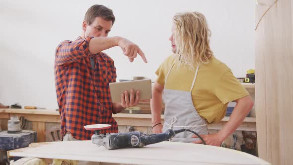 Two Caucasian male surfboard makers standing and working on projects using a tablet