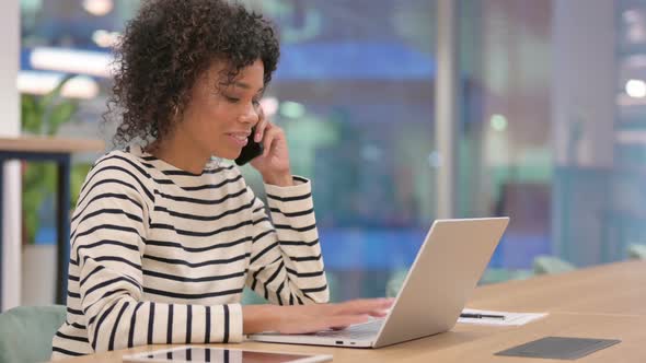 African Woman with Laptop Talking on Smartphone in Office