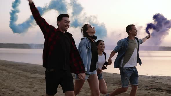 Young Four People Running with Colored Smoke Grenade in Their Hands By the Sea During Sunset