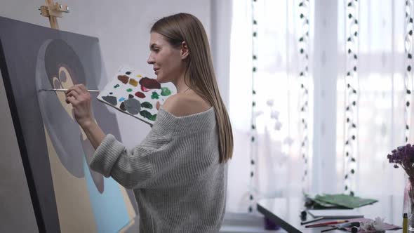 Talented Cheerful Young Woman Painting on Canvas Waving Looking Away Smiling Inviting Guest