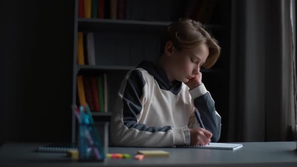 Portrait of Focused Pupil Boy Making Notes in Copybook with Pen Sitting at Desk in Dark Room