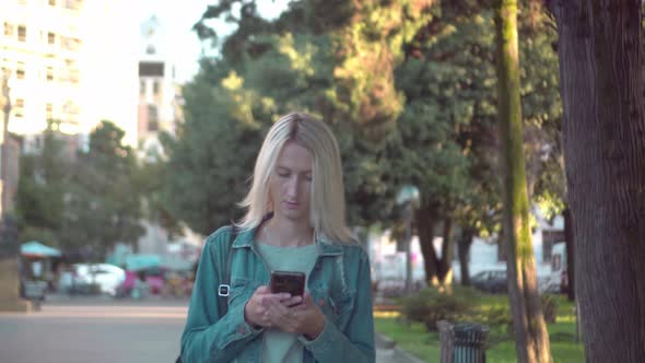 a blonde in a denim jacket walks around the city looking at her smartphone