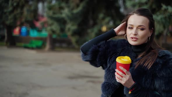 Fashionable girl in the park. Portrait of a lovely woman in fur coat with plastic cup of coffee