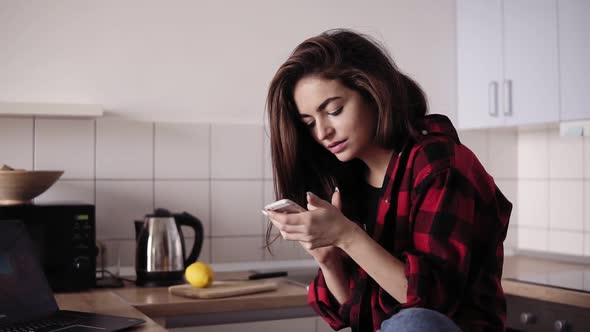 Young Beautiful Brunette Girl with Messy Hair in Flannel Shirt Sitting in Her Kitchen and Texting