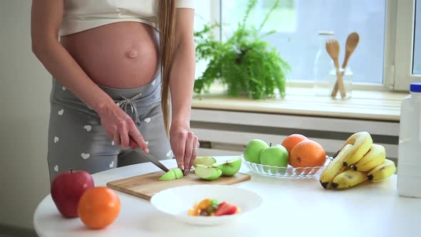 Pregnant Woman Cutting Fresh Apple and Standing at Table in Home Kitchen Spbd