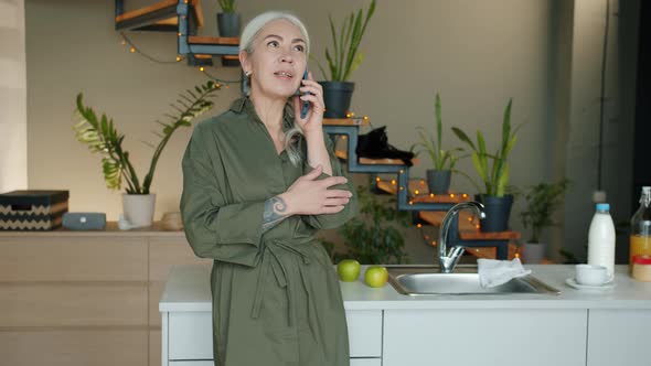 Cheerful Mature Woman Speaking on Mobile Phone and Smiling Standing Indoors in Kitchen