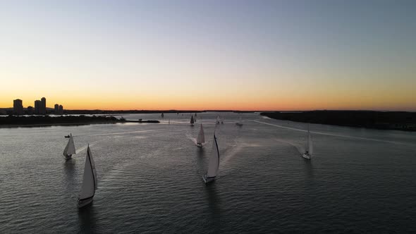 A group ofing sailing boats racing along the calm waters of a city harbor against a colorful sunset