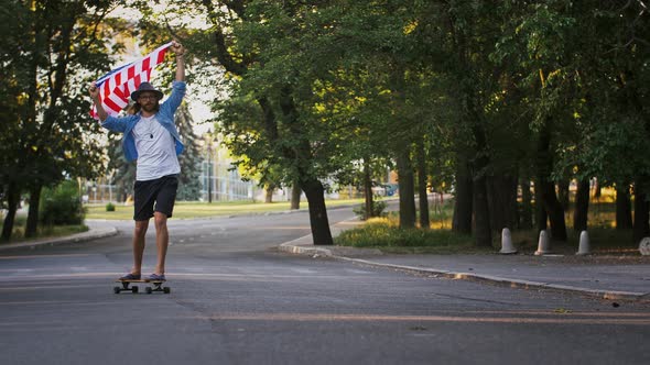 Stylish Young Guy Holding Waving Flag of USA Above His Head Hanging It on Neck Riding Skateboard