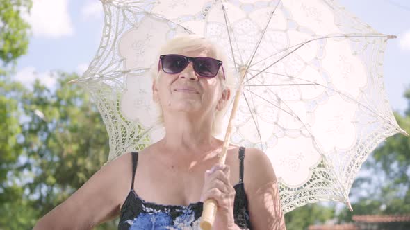 Portrait of Positive Smiling Mature Woman in Sunglasses Standing in the Park Under the White Parasol