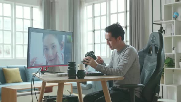 Asian Cameraman Holding And Looking At Camera While Using Desktop Computer For Editing Photo At Home