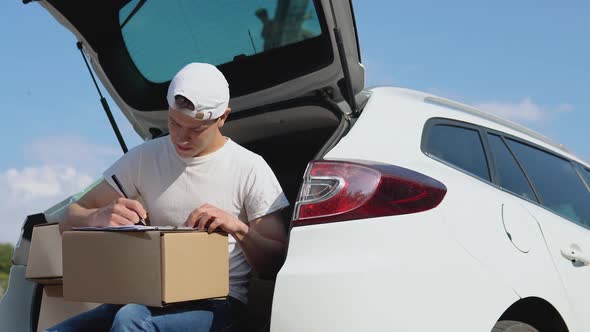 The Courier Sits on the Edge of the Open Trunk Filled with Boxes of Goods and Fills Out Documents