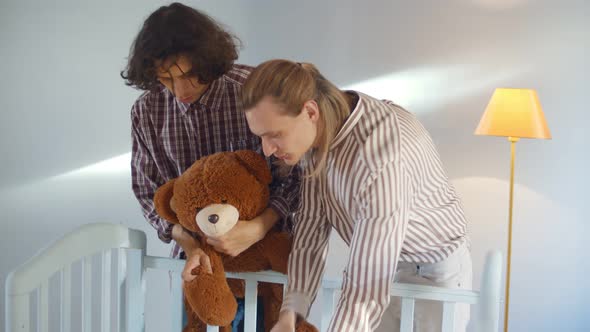 Homosexual Male Couple Playing with Adopted Baby in Crib Holding Teddy Bear