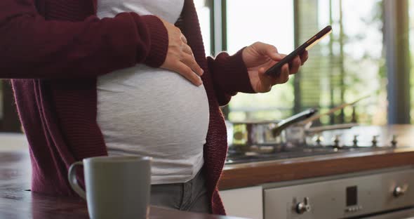Midsection of caucasian pregnant woman touching belly and using smartphone