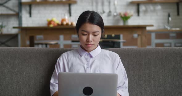 Asian Woman Resting on Couch Using Laptop Notebook Looking at Screen Typing Message at Living Room