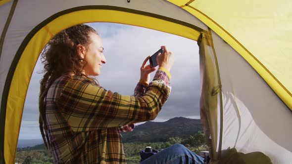 Happy caucasian woman camping, sitting outside tent on rural mountain taking photo with smartphone