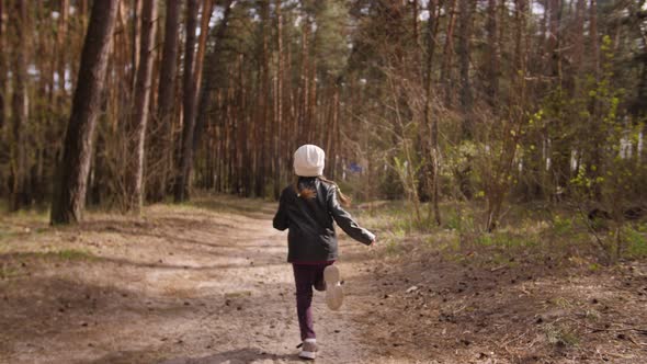 The Little Girl Runs Along the Path Road in the Pine Forest. Dressed in the Leather Jacket.