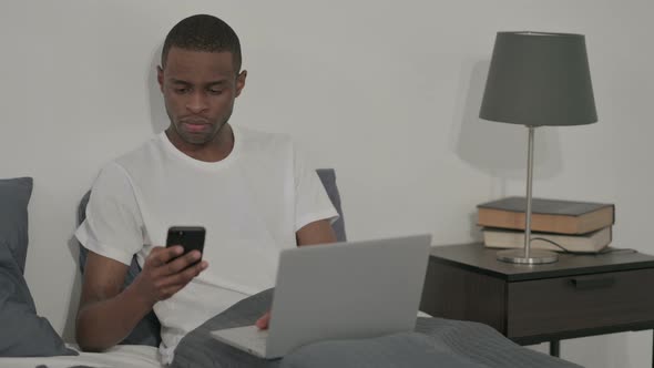 African Man with Laptop Using Smartphone Sitting in Bed