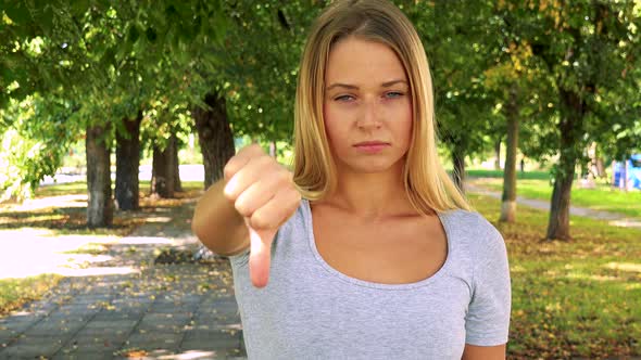 Young Pretty Blond Woman Disagrees (Show Thumb Down) - Park with Trees in Background