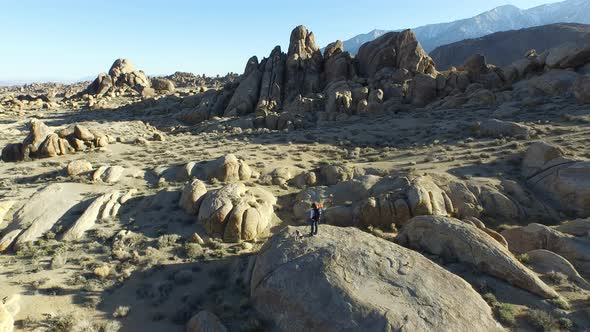 Aerial shot of a young man backpacker standing on a boulder with his dog in a desert mountain range.