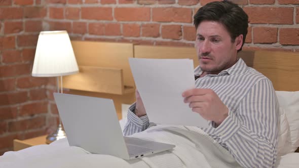 Middle Aged Man with Laptop having Loss on Documents in Bed