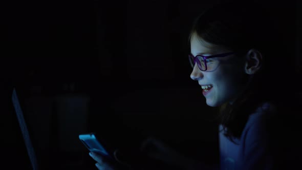Girl in eyeglasses playing on smartphone and laptop at night alt