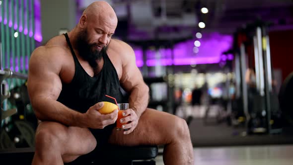 A Male Bodybuilder with Fruit Juice Relaxes Enjoying After a Workout Sitting on the Simulator in the