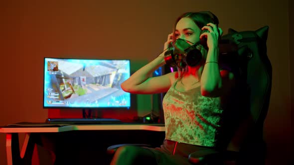 Young Gamer Woman in a Black Respirator Sitting By the PCand Putting on Big Headphones