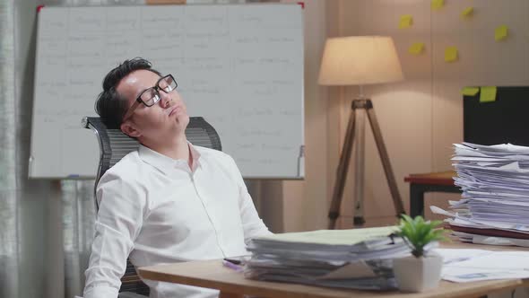 Tired Asian Man Leaning On The Office Chair And Sleeping While Working Hard With Documents