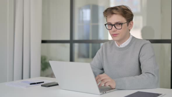 Young Man Smiling at Camera While Using Laptop in Modern Office