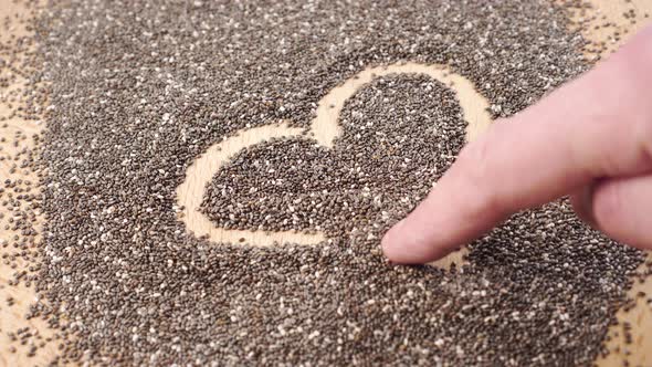 Hand is drawing a heart symbol on a heap of chia seeds. 