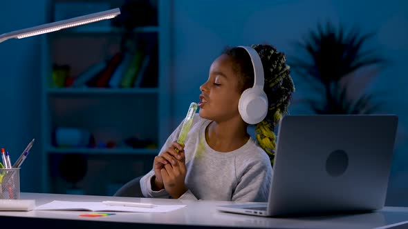 A Girl in White Headphones Sitting at a Desk Under a Table Lamp Listens to Music From Her Laptop