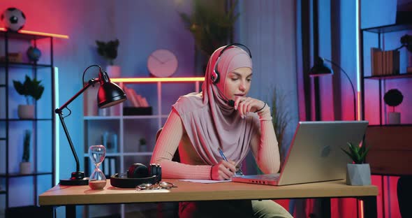 Muslim Woman in Hijab Wears Headphones E-learning while Listening Online Webinar at Home