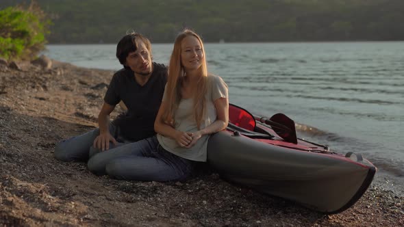 A Young Man and Woman Tourists Sitting By a Big Inflatable Kayak on a Seashore or a Lake