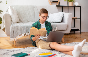 Smart school boy writing essay, holding book and using laptop