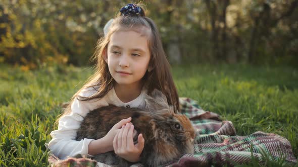 Little Girl Hugging and Stroking a Fluffy Brown Rabbit on Green Grass.