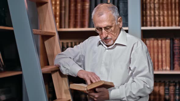 Senior Male Scientist Professor Teacher Reading Antique Book Standing on Stairs at Public Library