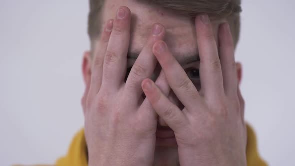 Close-up of a Shy Young Man Looking at the Camera From Behind Fingers Covering Face with His Hands