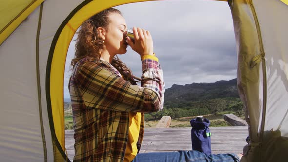 Happy caucasian woman camping, sitting outside tent drinking coffee in rural mountain setting