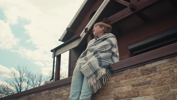 An adult caucasian woman sits covered with a plaid on the terrace of a country house