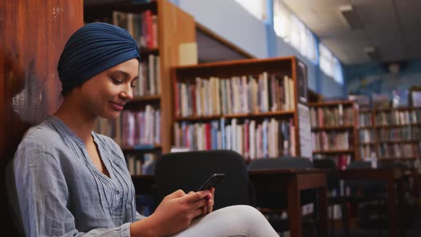 A happy Asian female student wearing a blue hijab sitting and using a smartphone