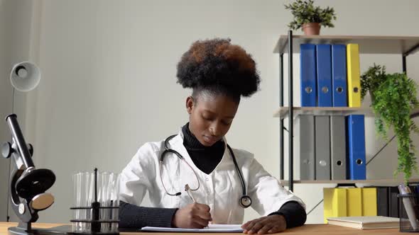 African American Female Doctor Writing Medical History of Patient While Sitting at Table in Hospital