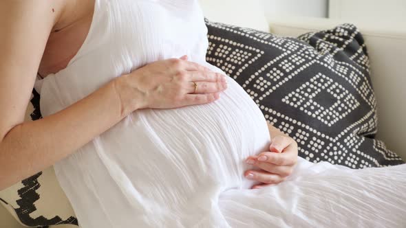 Female Hands Strokes Belly Sitting on Sofa with Soft Pillows in Light Room at Home